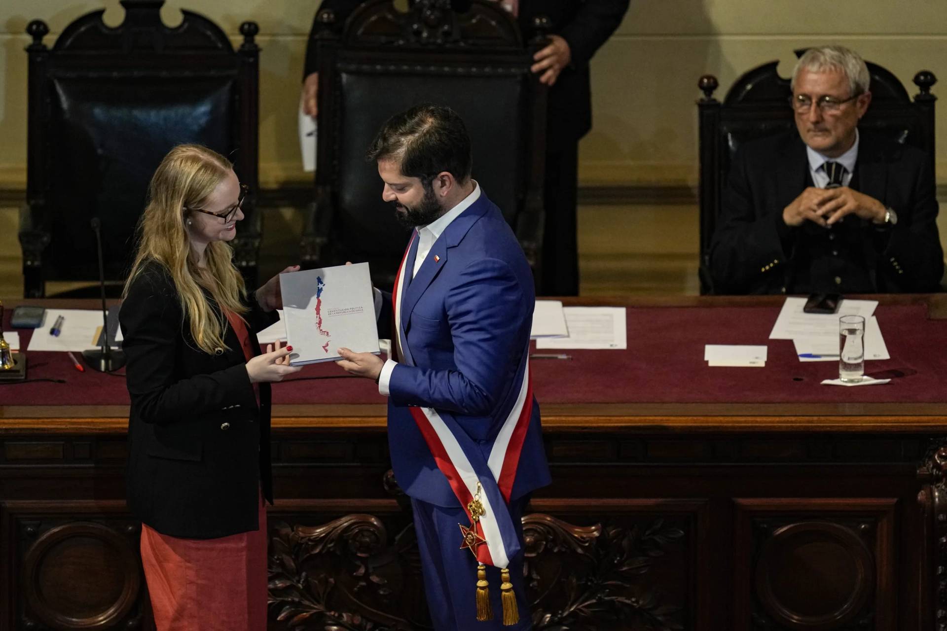Constitutional Council President Breatriz Hevia, left, presents Chilean President Gabriel Boric with the final draft of the new national Constitution by as Constitutional Council Vice President Aldo Valle looks on during a ceremony at the former National Congress building in Santiago, Chile, Tuesday, Nov. 7, 2023. (Credit: Esteban Felix/AP.)