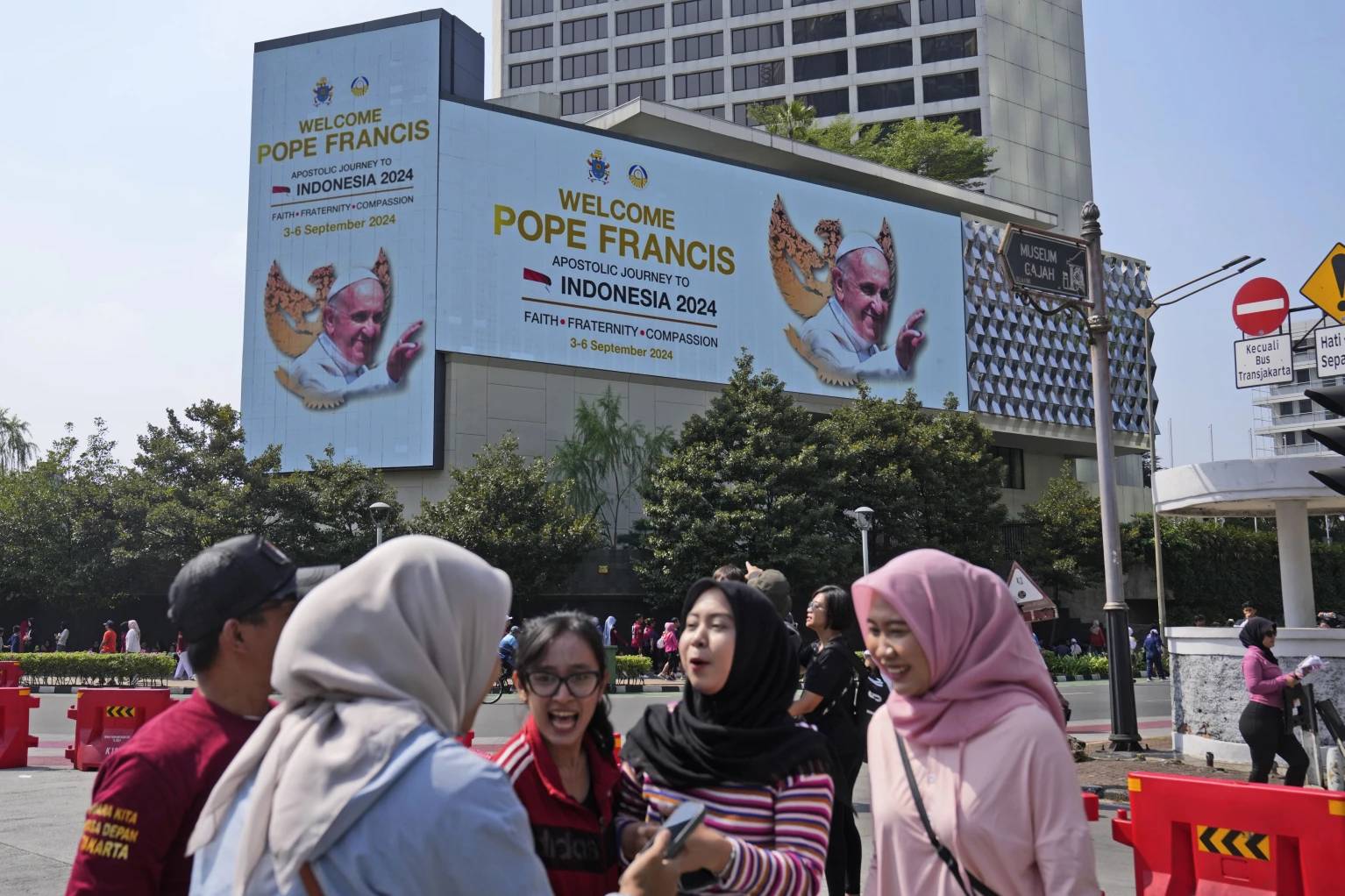 Muslim women chat as a large digital advertisement board with a welcoming message for Pope Francis is displayed on the facade of a building nearby in Jakarta, Sunday, Sept. 1, 2024, ahead of his visit to Indonesia from Sept. 3-6. (Credit: Dita Alangkara/AP.)