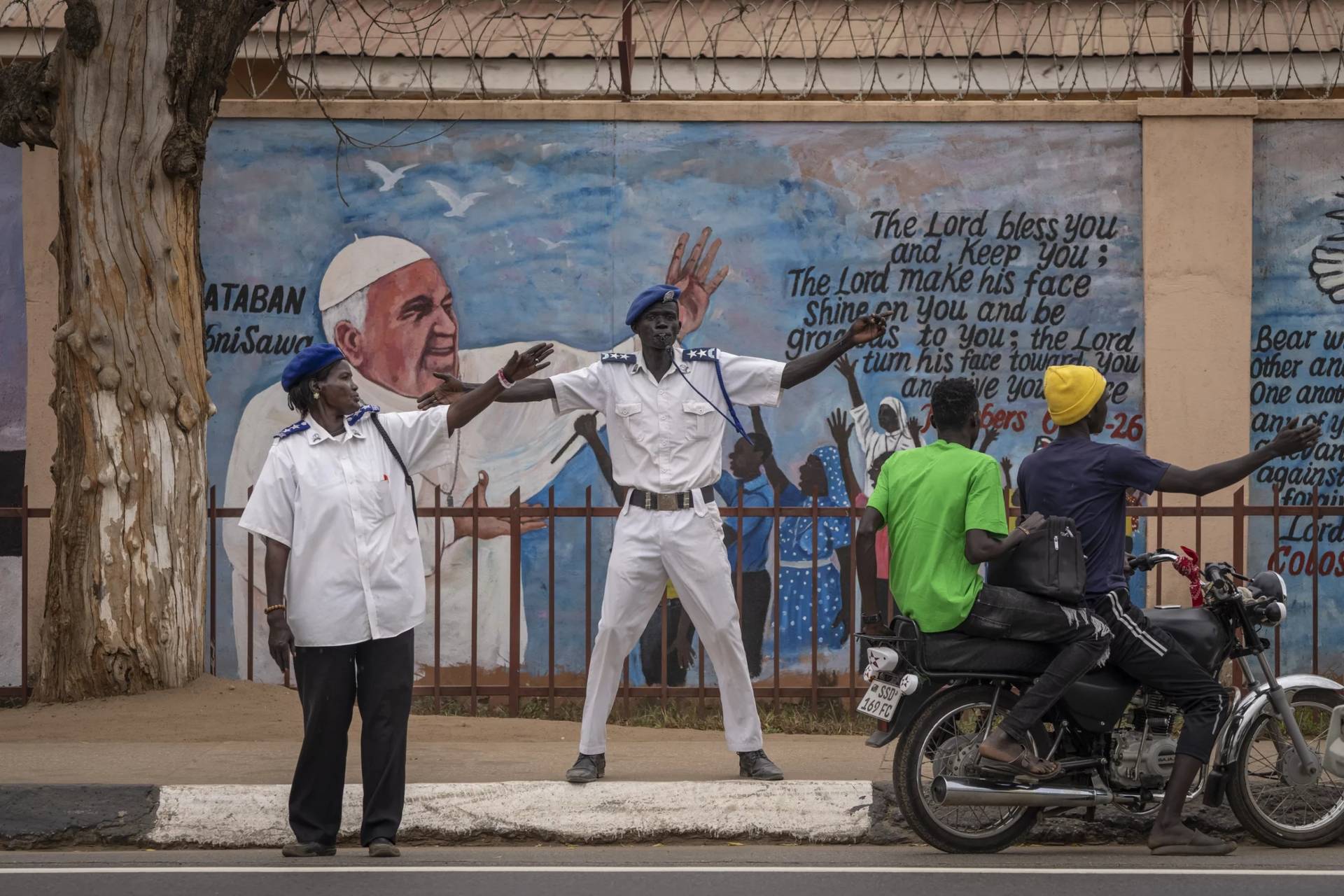 Standing by a mural of Pope Francis on a church, traffic police direct vehicles to get off the road as a government vehicle convoy passes through Juba, South Sudan, Feb. 2, 2023. (Credit: Ben Curtis/AP.)