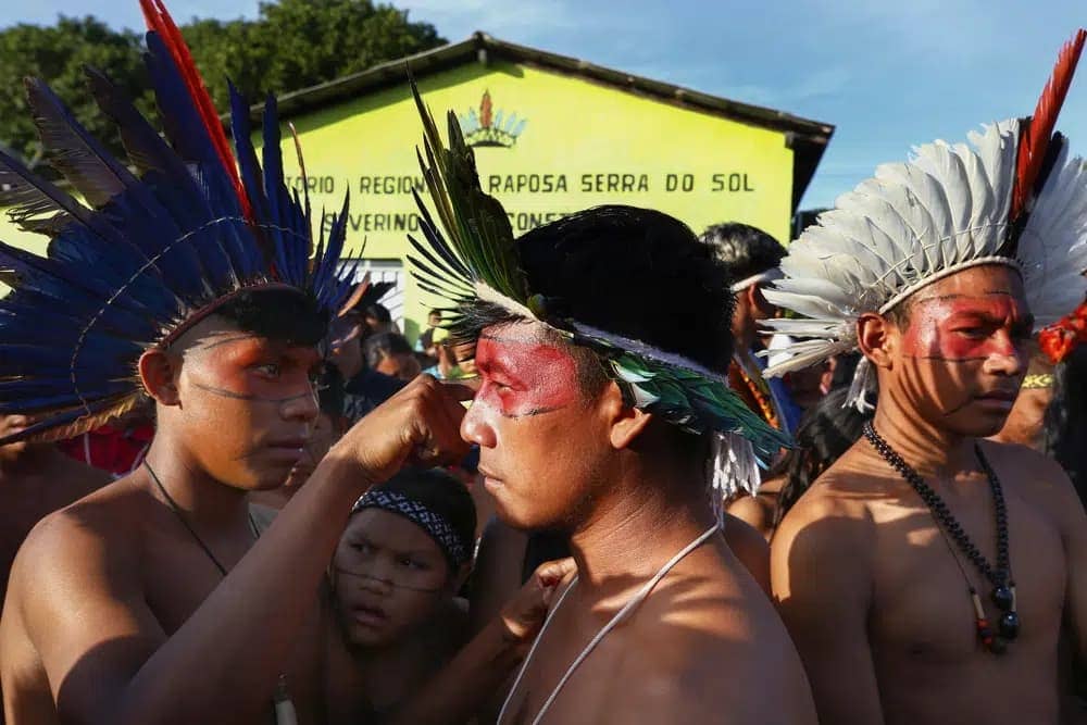 A Macuxi Indigenous man paints the face of another at the Caracarana Lake Regional Center in Normandia, on the Raposa Serra do Sol Indigenous reserve in Roraima state, Brazil, Monday, March 13, 2023. (Credit: Edmar Barros/AP.)