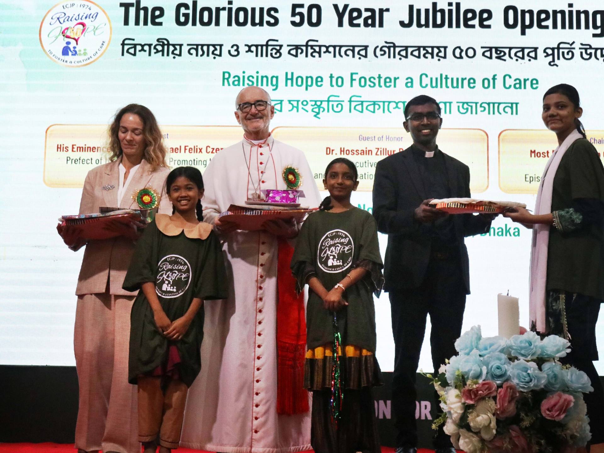 Cardinal Michael Felix Czerny (middle), Francesca Dona (right), regional coordinator-Asia Mainland (left), Father Joseph Savarimathu (right), taking a gift souvenir from the children. Children also provide a gift to Pope Leo through Cardinal Czerny. (Credit: Stephan Uttom Rozario.)