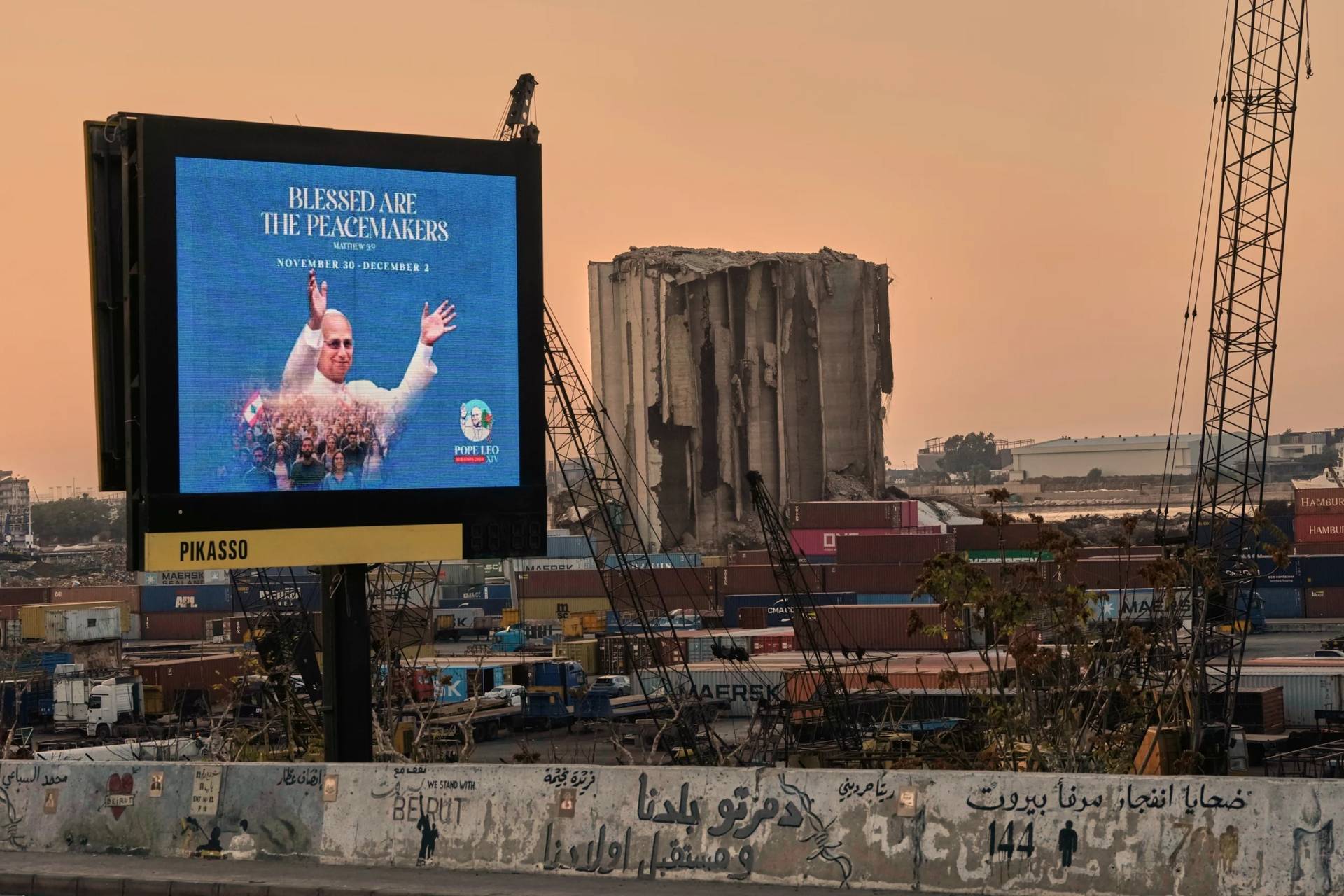 A billboard displays an image of Pope Leo XIV, ahead of his upcoming visit to Lebanon, near the grain silos at Beirut's port that were heavily damaged in the massive 2020 explosion as a graffiti in Arabic reading "Victims of the Beirut port explosion" is seen in the foreground, Thursday, Nov. 20, 2025. (AP Photo/Hassan Ammar)