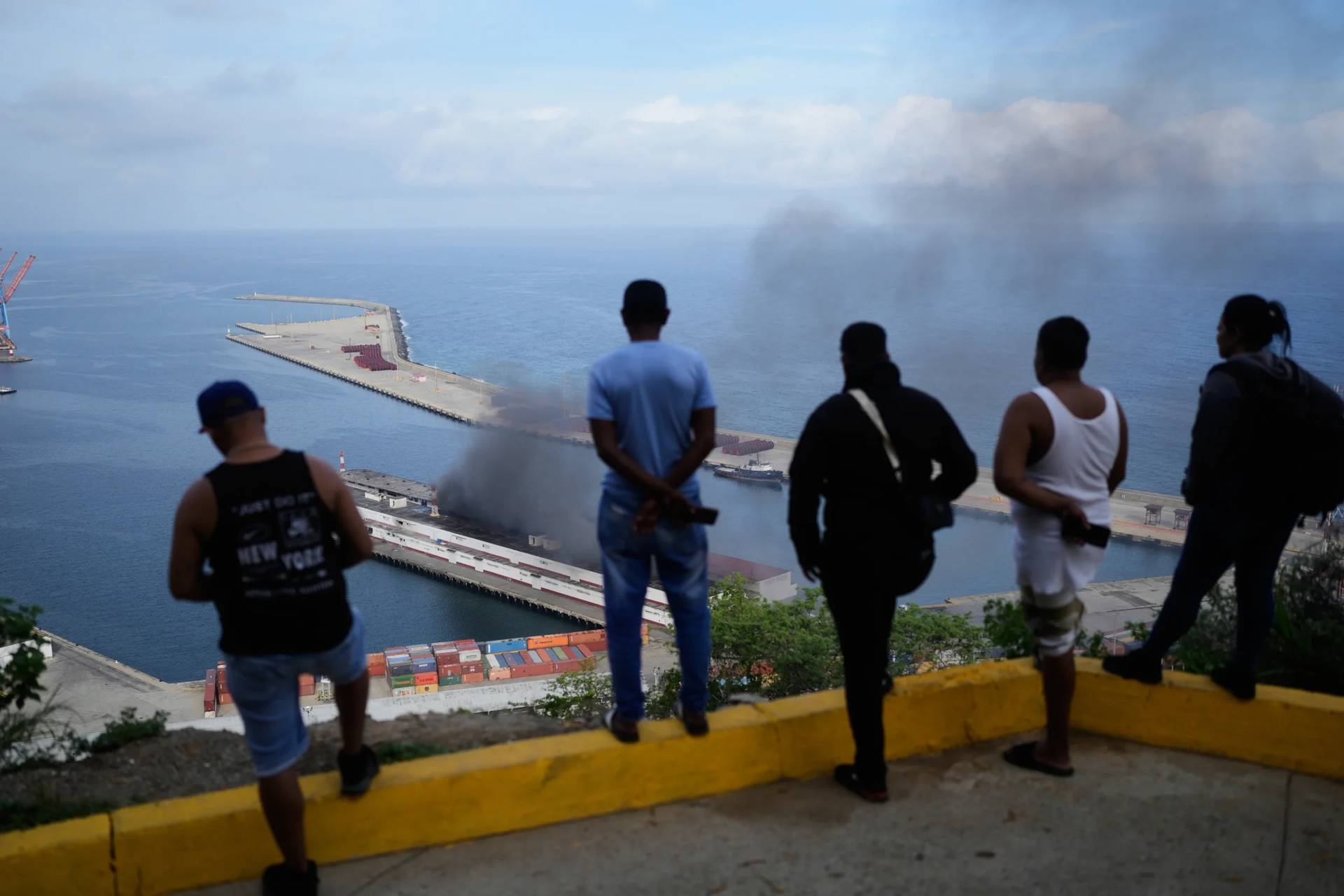 Men watch smoke rising from a dock after explosions were heard at La Guaira port, Venezuela, Saturday, Jan. 3, 2026. (Credit: Matias Delacroix/AP.)