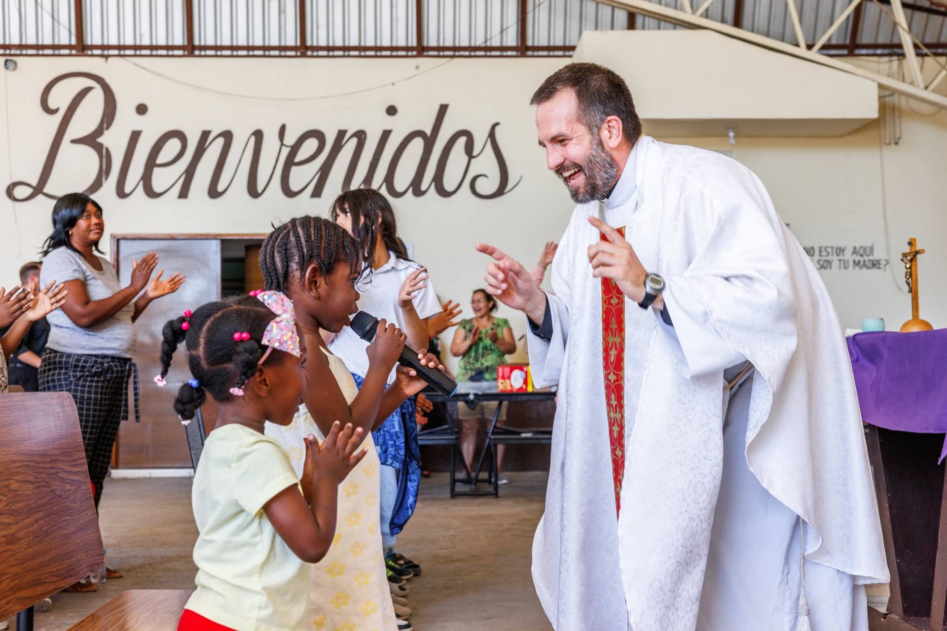 Father Brian Strassburger smiles as Alcala Bouilly sings into the microphone during Mass at Casa del Migrante on Thursday, March 19, 2026, in Reynosa, Mexico. (Credit: Michael Gonzalez/AP.)