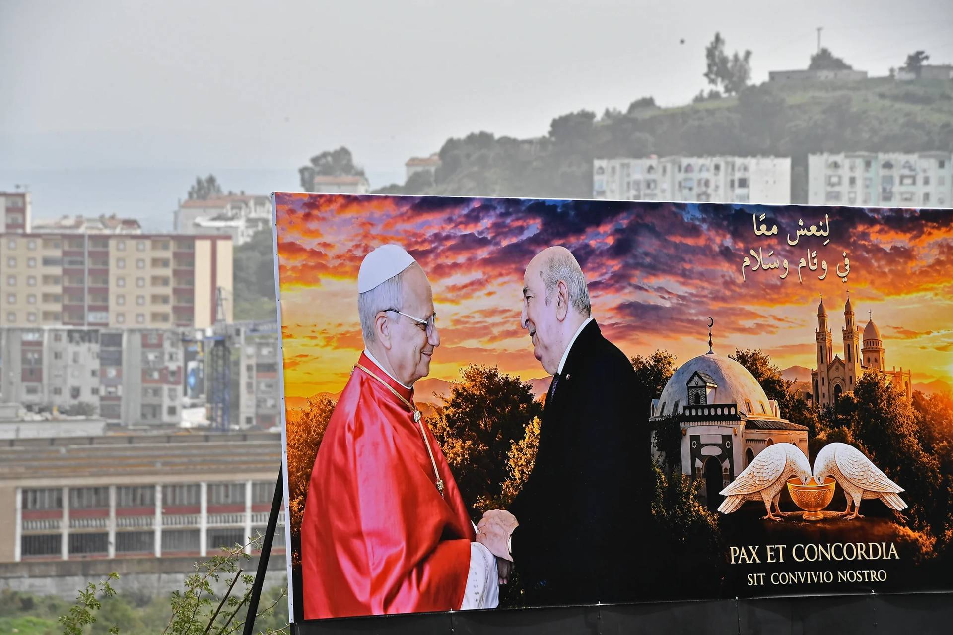 A banner showing a photo of Pope Leo XIV and Algerian President Abdelmadjid Tebboune in Annaba, eastern Algeria, Saturday, April 11, 2026, ahead of Pope Leo XIV’s visit. The banner in Arabic reads, “Let’s live in peace and harmony.” (Credit: Fateh Guidoum/AP.)