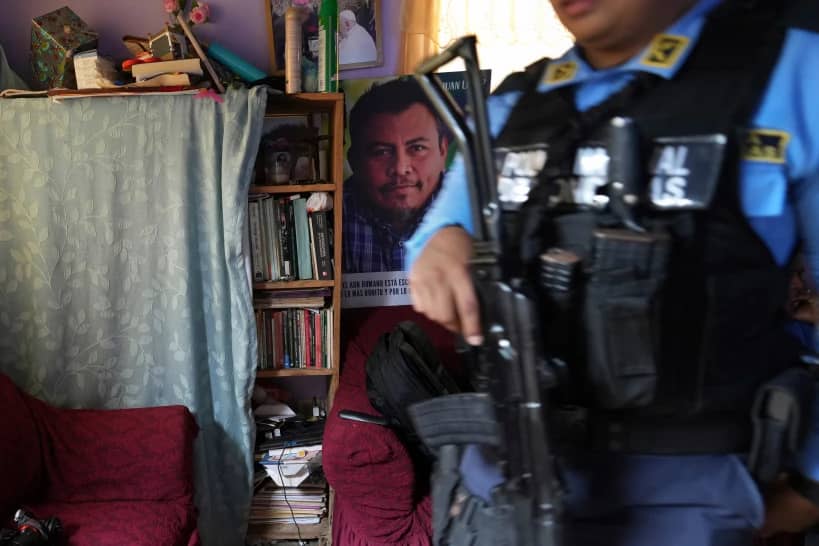 A poster of slain environmentalist Juan López hangs in his home as a state-assigned bodyguard watches over the López family, in Tocoa, Honduras, Dec. 6, 2024. (Credit: Moises Castillo/AP.)